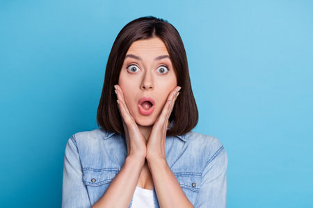 Portrait of impressed young lady hands on cheeks open mouth look camera isolated on blue color...
