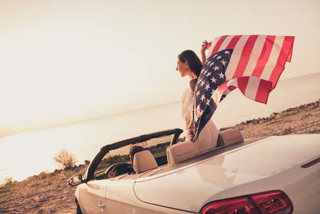 Photo of funny carefree married couple dressed white clothes driving car holding american flag outdoors country side roadの写真素材