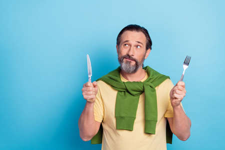 Photo of handsome minded man hold knife fork look interested empty space isolated on blue color backgroundの写真素材