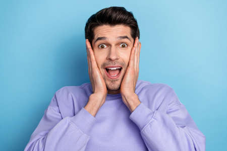 Photo of impressed young brunet guy hands face wear sweatshirt isolated on blue color background