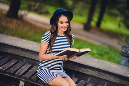 Portrait of attractive cheerful focused girl sitting on bench reading interesting academic book on fresh air outdoorsの写真素材