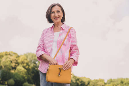 Photo of adorable sweet retired woman dressed striped shirt walking holding purse outside...