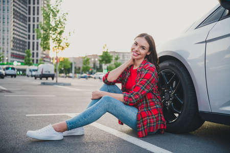 Portrait of attractive cheerful girl driver sitting on floor near new modern car ownership dealer on...