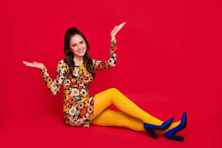 Photo of sweet pretty woman wear flower print dress sitting floor shrugging shoulders isolated red color backgroundの写真素材