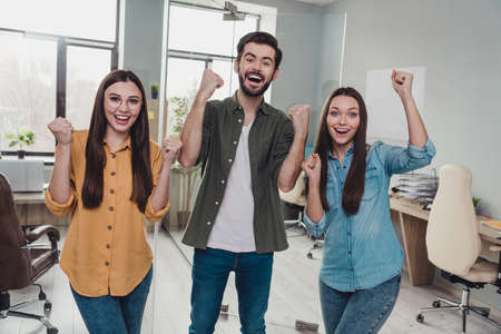 Portrait of three delighted employees raise fists celebrate attainment shout yes office indoors