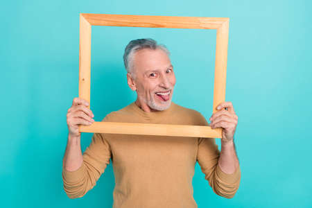 Photo of attractive good mood male fooling around stick out tongue holding wooden frame isolated on teal color backgroundの写真素材