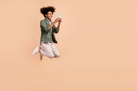 Full length body size view of attractive cheerful guy jumping using device chatting isolated over beige pastel color backgroundの写真素材