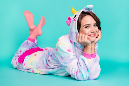 Photo of shiny dreamy young woman dressed unicorn nightwear smiling lying floor arms cheeks isolated teal color backgroundの写真素材