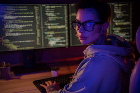 Portrait of intelligent hipster hacker girl sitting chair keyboard typing operating script dark room indoorsの写真素材
