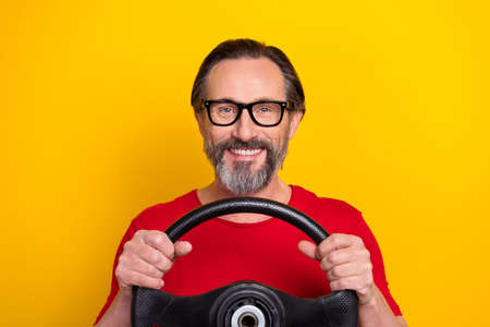 Photo of cheerful pretty retired man wear red t-shirt spectacles riding car smiling isolated yellow...