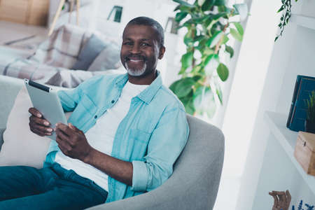 Photo of positive cheerful man sitting chair hold use tablet toothy smile good mood free time house indoorsの写真素材