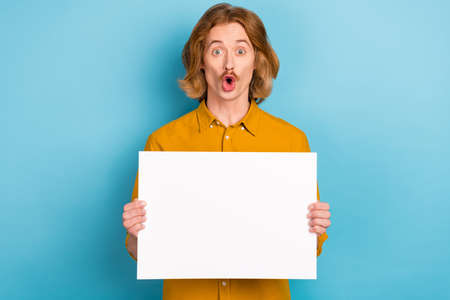 Portrait of attractive trendy stunned long-haired guy holding copy space banner isolated over bright...