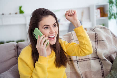 Portrait of attractive trendy cheerful girl sitting on divan talking on phone rejoicing good news at...