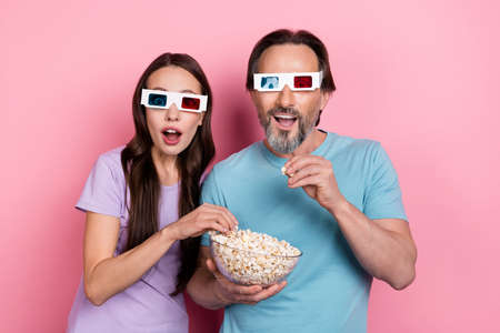 Photo of two shocked surprised funky people eat popcorn watch movie 3d glasses isolated on pink...