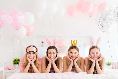 Portrait of four adorable girls hands touch cheeks laying white sheets bed house indoorsの写真素材