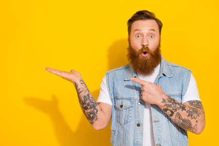 Photo of funny impressed guy dressed denim vest holding arm pointing empty space isolated yellow...