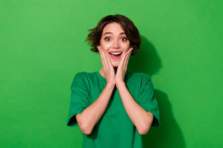 Photo of astonished cheerful girl arms touch cheeks look camera isolated on green color background