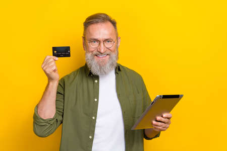 Portrait of positive aged man hold debit plastic card tablet toothy smile isolated on yellow color...