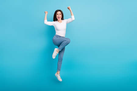 Photo of funny lucky woman wear white shirt rising fists jumping high empty space isolated blue...