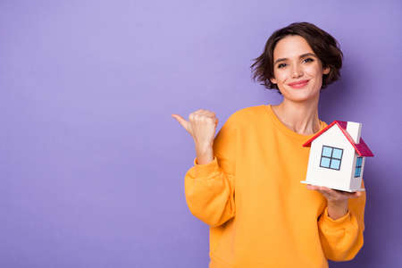 Photo of attractive positive lady indicate thumb finger empty space hold little house isolated on violet color backgroundの写真素材