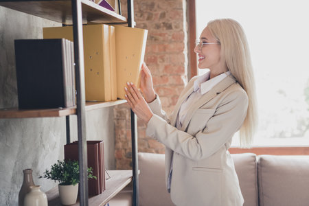 Profile portrait of cheerful charming lady stand bookshelf take documents folder modern loft office insideの写真素材