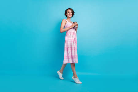 Full body photo of cute millennial lady go drink tea wear pink long sarafan sneakers isolated on blue backgroundの写真素材