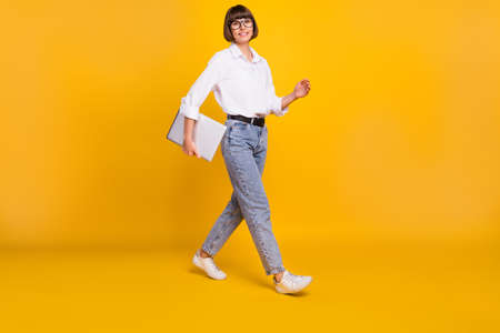 Photo of positive geek lady carry netbook walk wear eyewear white shirt jeans shoes isolated yellow color backgroundの写真素材