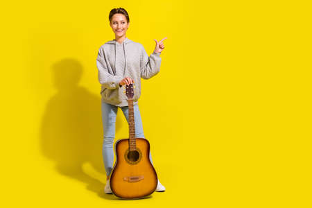 Full length photo of funny brown hair millennial lady with guitar point empty space wear sweater jeans sneakers isolated on yellow backgroundの写真素材