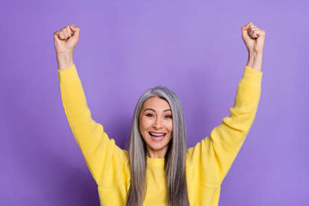 Photo of cheerful laughing mature female raise fists in victory win lottery prize isolated on purple color backgroundの写真素材