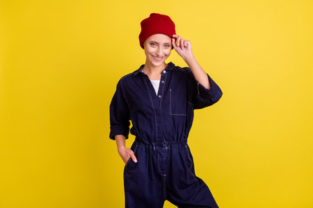 Photo of young beautiful smiling confident hipster woman wear denim overall red beanie isolated on yellow color backgroundの写真素材
