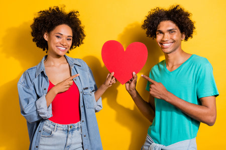 Photo of happy dark skin man and woman people couple point finger heart isolated on yellow color backgroundの写真素材