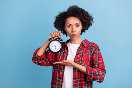 Photo of young black girl amazed shocked demonstrate clock time deadline isolated over blue color backgroundの写真素材
