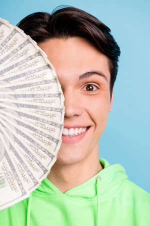 Vertical photo of cheerful guy hold cash fan cover half face wear green sweatshirt isolated blue color backgroundの写真素材