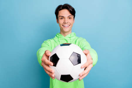 Photo of excited sweet young guy dressed green hoodie inviting play football smiling isolated blue...