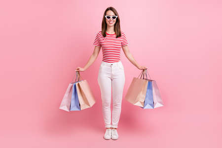 Full body photo of sweet young brunette lady hold bags wear eyewear striped t-shirt jeans isolated on pink color backgroundの写真素材