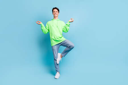Photo of peaceful dreamy guy meditate close eyes stand one leg wear green hoodie isolated blue color...