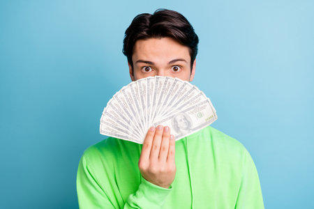 Photo of calm incognito rich guy hold cash fan close mouth wear green sweatshirt isolated blue color backgroundの写真素材