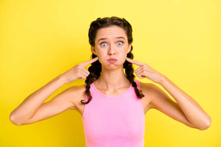 Photo of cute childish young woman pigtails wear tank top pointing fingers bloated cheeks isolated...