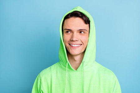 Photo of funny pretty young guy dressed green hoodie looking empty space smiling isolated blue color backgroundの写真素材