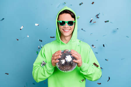 Photo of charming sweet young guy dressed green hoodie dark glasses holding disco ball smiling isolated blue color backgroundの写真素材