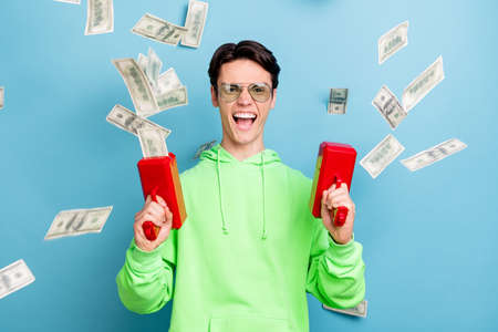 Photo of funky cool young guy dressed green hoodie shooting money gun smiling isolated blue color backgroundの写真素材