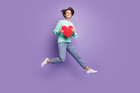 Full length body size photo of cheerful happy model jumping keeping red heart love symbol isolated pastel purple color backgroundの写真素材