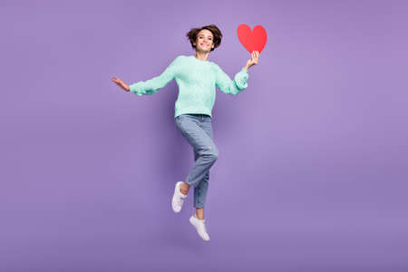 Full length body size view of attractive cheerful girl jumping holding red heart card isolated over violet purple color backgroundの写真素材