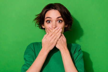 Photo of impressed worried girl arms palms cover face mouth look camera isolated on green color backgroundの写真素材
