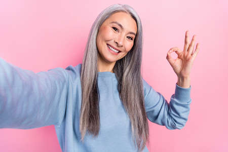 Photo of pretty cute retired woman blue sweater showing okey sign tacking selfie isolated pink color...