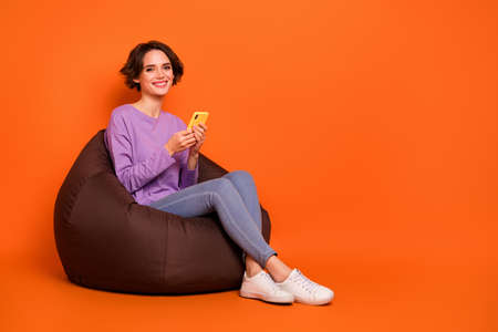 Full body portrait of cheerful nice lady sitting comfort bag use telephone isolated on orange color...