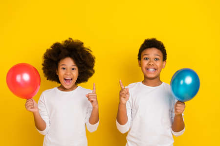 Portrait of two excited positive siblings hold air balloon point finger isolated on yellow color backgroundの写真素材