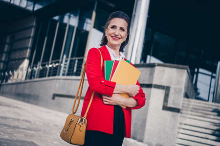 Photo of charming cheerful mature lady secretary wear red jacket holding documents folders outside city business complexの写真素材