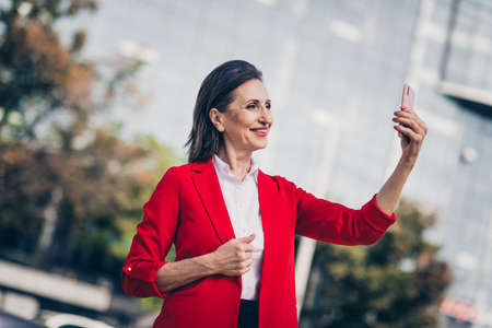 Photo of cute cheerful mature lady agent wear red jacket recording video modern gadget outside city...