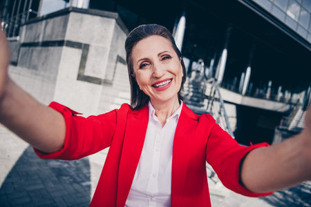 Photo of sweet pretty mature lady boss wear red jacket tacking selfie smiling outside city business complexの写真素材